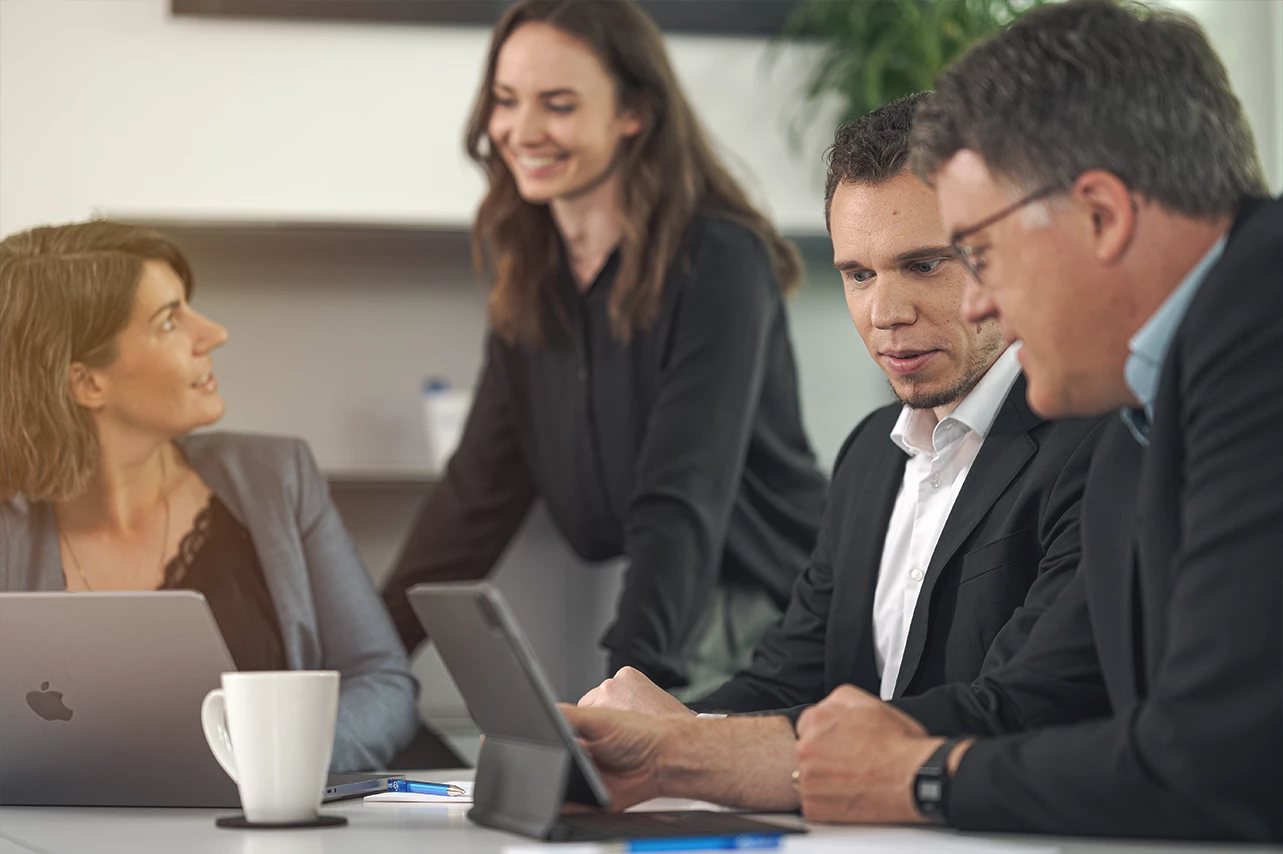 Vier SAP Berater in einem paricon Büro, die um einen Tisch sitzen und stehen, während sie an Laptops und Tablets arbeiten und miteinander sprechen.